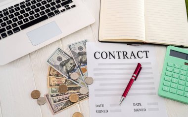 Basit bir masada dizüstü bilgisayar, dolar, bozuk para, sözleşmeli bir kalem ve bir defter var. İş konsepti