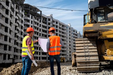 Tulum ve kask takmış iki işçi yeni binanın yanında dikilip konuşuyorlar. İnşaat konsepti