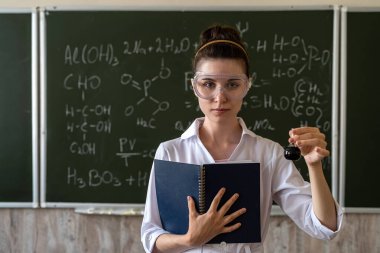 Kadın kimyager deney yapıyor ve renkli sıvılara bakıyor, sınıfta karatahtaya karşı.