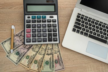 Tahta bir masada dizüstü bilgisayar, hesap makinesi, dolar banknotları, kalem var. İş konsepti