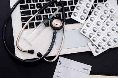 Siyah arka planda steteskop, hap, şırınga ve reçeteli bir laptop var. Tıp konsepti