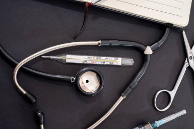 Siyah arka planda steteskop, burun damlası, şırınga, defter ve kalem var. Tıp konsepti