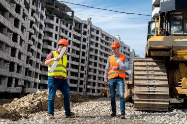 Üniformalı ve miğferli iki genç işçi inşaat sahasında bir derececinin yanında duruyor. İnşaat konsepti