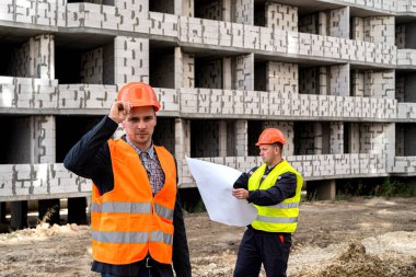 Tulumlu genç bir işçi elini kaskından tutuyor. Tulumlu başka bir işçi ise yeni bina için bir çalışma planı hazırlıyor. İnşaat konsepti