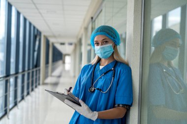 Genç ve güzel hemşire üniforma, maske, eldiven ve steteskop ile koridorda duruyor ve elinde bir tablet var. Tıp konsepti