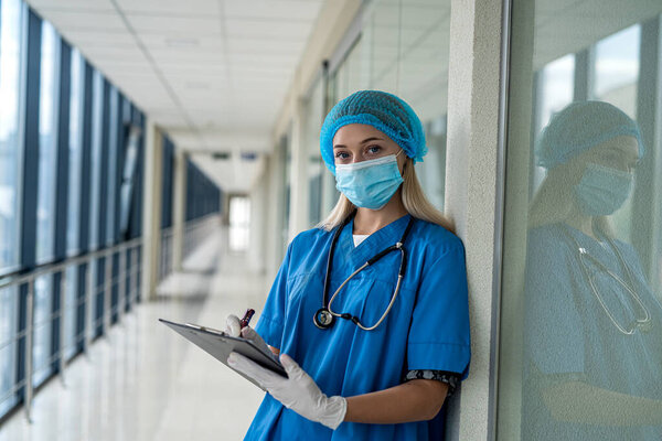 young beautiful nurse stands in the hallway in uniform, mask, gloves, stethoscope and holds a tablet. Medicine concept