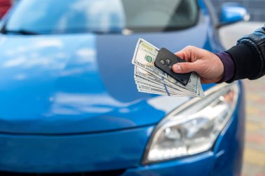 Erkek, mavi güzel bir arabanın arka planında dolar ve araba anahtarı tutan takım elbiseli bir adam. İş konsepti. Araba satın alma konsepti. İş konsepti