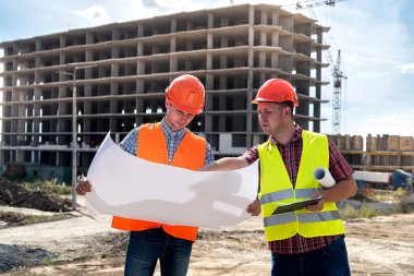 Üniformalı ve kasklı iki genç işçi, binaların arka planındaki ayrıntılı inşaat planına bakıyor. İnşaat konsepti
