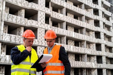 Üniformalı ve miğferli işçiler yeni binanın yakınındaki inşaat planını tabletle karşılaştırıyorlar. Yeni bir bina konsepti