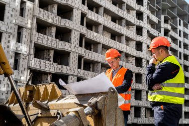 Üniformalı ve miğferli harika erkek işçiler yeni bir inşaatın planını bir not makinesine kağıt koyarak düşünürler. İnşaat konsepti