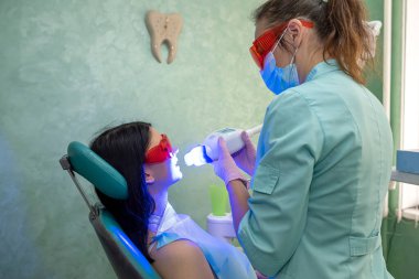 Dişçi koltuğunda oturan genç bir kadın doktorların kontrolü altında beyazlatma prosedürü uyguluyor.
