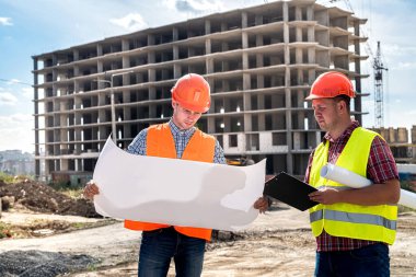 Üniformalı ve kasklı iki genç işçi, binaların arka planındaki ayrıntılı inşaat planına bakıyor. İnşaat konsepti