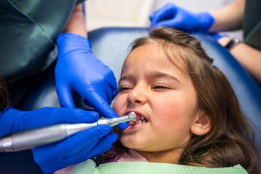 Özel kıyafetli genç dişçiler dişçi koltuğunda oturan küçük bir kızın dişlerini tedavi ediyorlar. Dişçilik ve çocuk kavramı