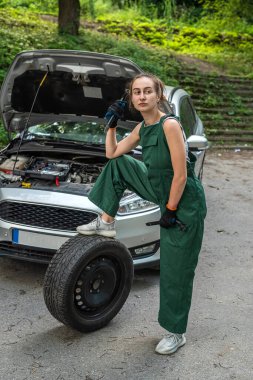 Yeşil üniformalı genç bayan tamirci bozuk bir arabanın yanında duruyor ve dışarıda yardım bekliyor.