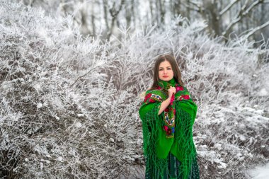 Yeşil elbiseli ve şallı güzel bir esmer kış ormanında duruyor..