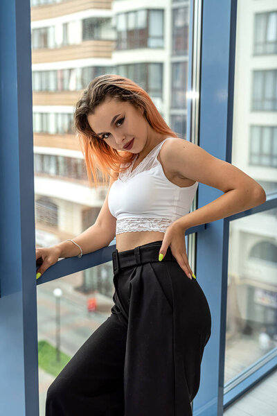 Sexy flexible perfect girl in dark black pants and white top curves near a spacious window. Beauty girl concept by the window