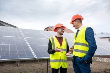 Miğferli ve yelekli patron güneş panellerinin yanında özel kıyafetler giyen bir çalışanın yanında duruyor. Yeşil elektrik kavramı