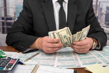 Geniş bir masada siyah şık takım elbiseli genç bir işadamı dolar sayıyor ve formları dolduruyor. Vergi formları ve dolar kavramı