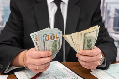Geniş bir masada siyah şık takım elbiseli yakışıklı bir genç adam dolar sayıp form dolduruyor. Vergi formları ve dolar kavramı