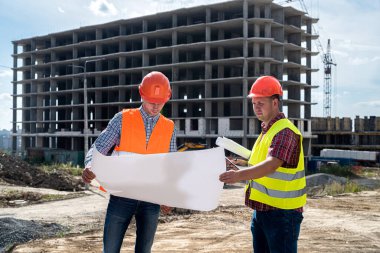 Üniformalı ve kasklı iki genç işçi, binaların arka planındaki ayrıntılı inşaat planına bakıyor. İnşaat konsepti