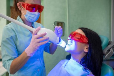 Kadın dişçi, dişçi hastasını ultraviyole lamba kullanarak dolduruyor. Diş beyazlatma prosedürü