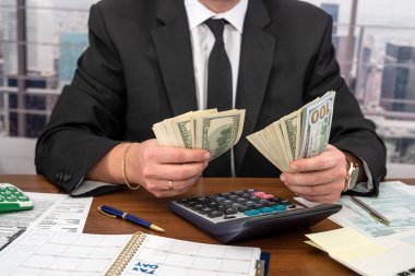 Geniş bir masada siyah şık takım elbiseli yakışıklı bir genç adam dolar sayıp form dolduruyor. Vergi formları ve dolar kavramı