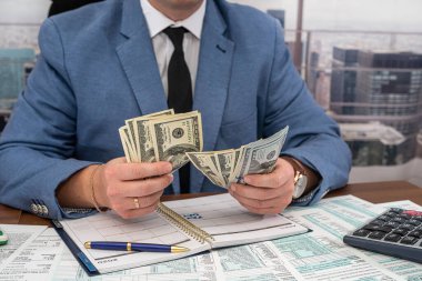 Geniş bir masada siyah şık takım elbiseli genç bir işadamı dolar sayıyor ve formları dolduruyor. Vergi formları ve dolar kavramı