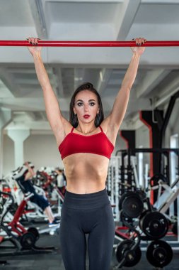 Esmer, kırmızı bluzlu, sıska taytlı, iyi fiziksel formda, esneme ve egzersiz yapan bir kız. Koridorda eşofman konsepti