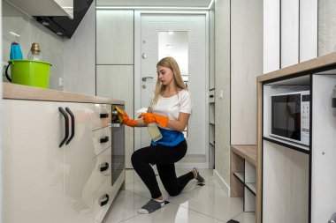 Genç, ince, renkli eldivenli, paçavra ve deterjanla mutfakta temizlik yapıyor. Mutfaktaki kızı temizleme fikri.
