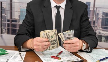 Geniş bir masada siyah şık takım elbiseli yakışıklı bir genç adam dolar sayıp form dolduruyor. Vergi formları ve dolar kavramı