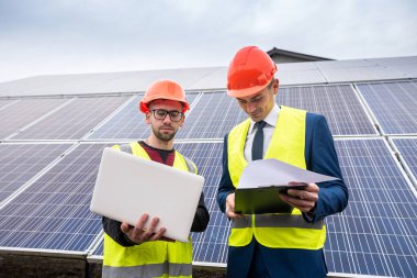 Üniformalı ve miğferli ustabaşı işadamına güneş panelleri kurma planını anlatıyor. Yeşil elektrik kavramı