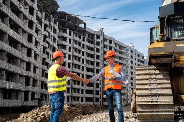 Üniformalı ve miğferli iki iyi işçi yeni bina ve greyderin yanında el sıkışıyor. İnşaat konsepti