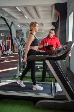Koşu bandında koşan kişisel antrenör yardımcısı olan bir bayan sporcu. Spor salonunda insanlar spor yapıyor.