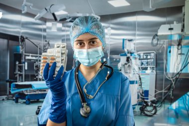 Genç, güzel bayan doktor mavi ceketli, steteskop tutan ve elinde haplarla hastanedeki ameliyathanede ayakta duran.