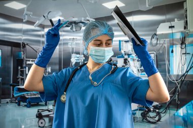 Mavi üniformalı ve koruyucu maskeli kadın doktor. Eldivenler hastanedeki ameliyat odasındaki panoya notlar yazıyor.