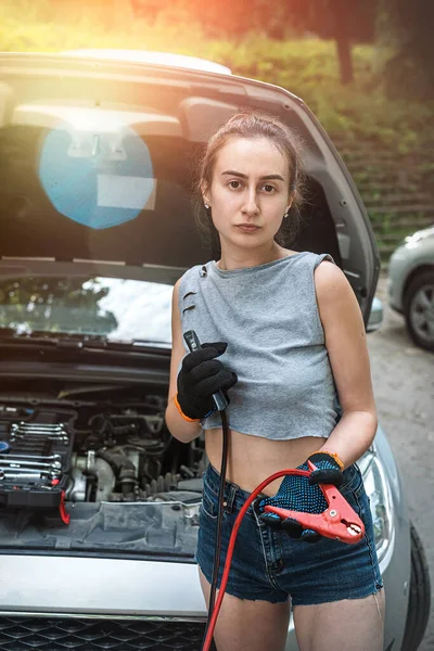 woman stand near broken car and holding black and red jumper cable for recharge the battery car. repair auto at summer trip