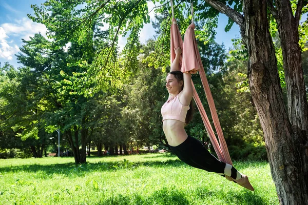 young strong woman with a thin waist with strong arms engaged in exercises on an air hammock in nature. Outdoor fly yoga concept