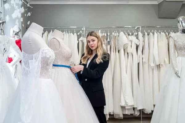 female tailor measuring elegant wedding dress on mannequin using tape measure in store