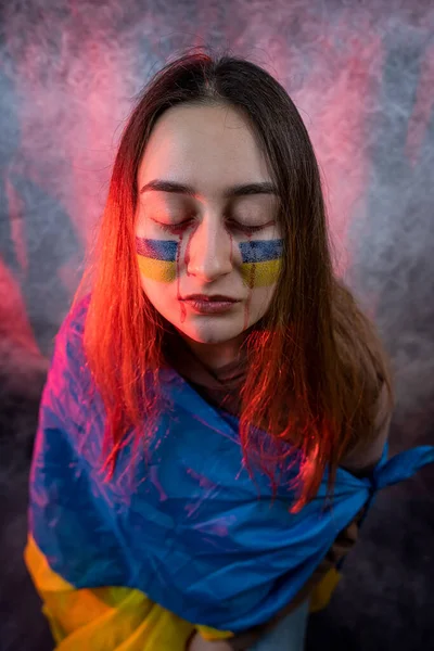 Pretty young frightened woman with Ukraine flag on face isolated on dard background. Concept peace. Pray for Ukraine