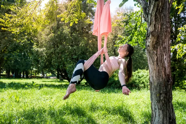 young trainer with gorgeous hair from fly yoga is engaged in stretching on an air hammock attached to a tree. Yoga fly concept on air