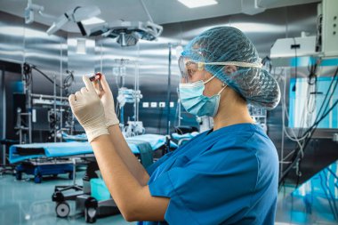 Üniformalı maske takan çekici ve deneyimli bir doktor. Eldiven takıyor. Hastanedeki ameliyathanede kan sondalarını inceliyor.