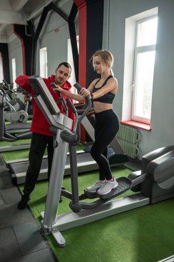 Koşu bandında koşan kişisel antrenör yardımcısı olan bir bayan sporcu. Spor salonunda insanlar spor yapıyor.