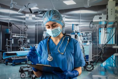 Mavi üniformalı ve koruyucu maskeli kadın doktor. Eldivenler hastanedeki ameliyat odasındaki panoya notlar yazıyor.