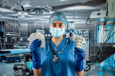 Genç, güzel bayan doktor mavi ceketli, steteskop tutan ve elinde haplarla hastanedeki ameliyathanede ayakta duran.