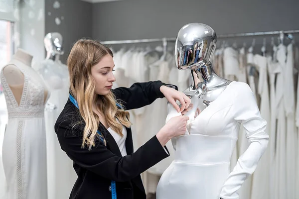 female tailor measuring elegant wedding dress on mannequin using tape measure in store