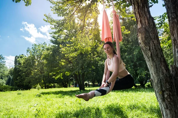 Happy physically developed woman practices yoga on aerial canvases on a tree. Outdoor yoga concept on tree