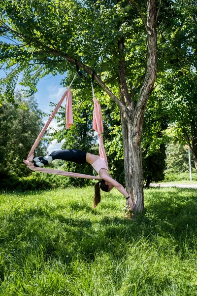 young trainer with gorgeous hair from fly yoga is engaged in stretching on an air hammock attached to a tree. Yoga fly concept on air