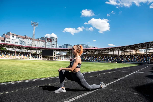 Spor giyimli genç ve zayıf bir kadın iş gününden önce stadyumda esneme hareketleri yapıyor.