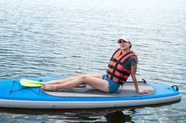 smiling woman sitting on sup board with paddle. resting and sunbathing on a nature
