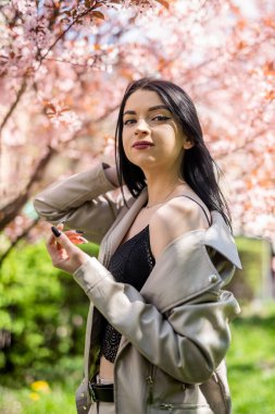 Beyaz deri ceketli şık bir kadın bahar bahçesinde kiraz çiçeğinin yanında poz veriyor.